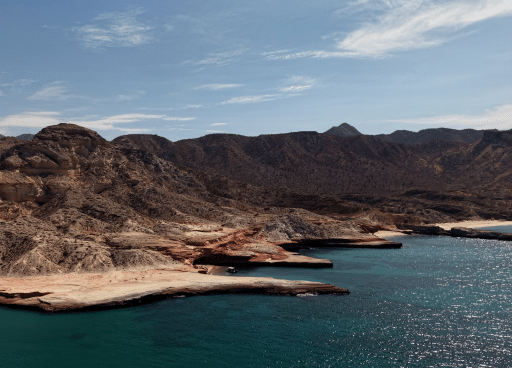 Aerial view of Isla San José's red sandstone coastline meeting the turquoise Sea of Cortez, desert mountains behind
